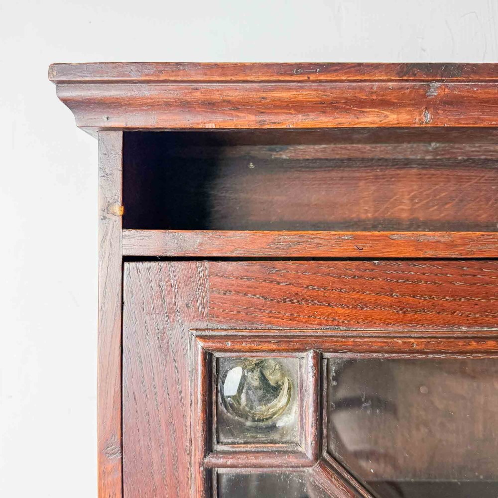 A dark oak wall-mounted cabinet with a single glazed door featuring X-shaped glazing bars, brass drop handle, open top shelf, and shaped hanging brackets. View XI