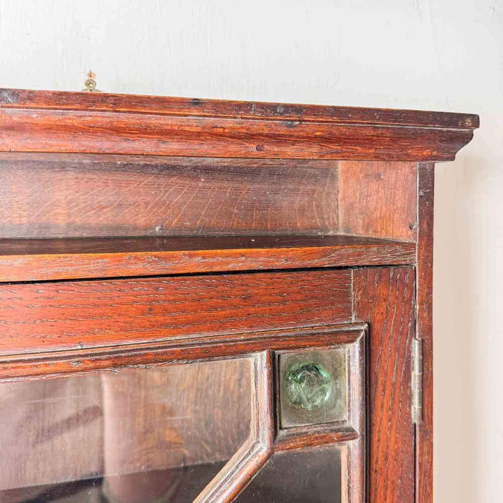A dark oak wall-mounted cabinet with a single glazed door featuring X-shaped glazing bars, brass drop handle, open top shelf, and shaped hanging brackets. View XIII