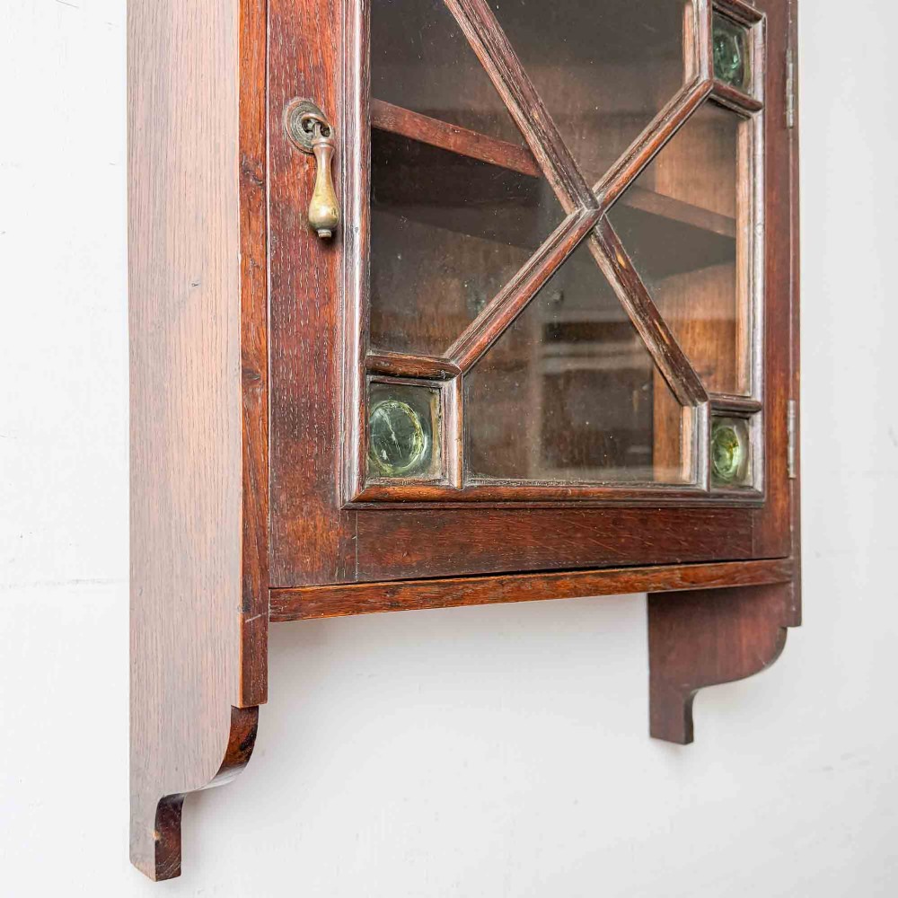 A dark oak wall-mounted cabinet with a single glazed door featuring X-shaped glazing bars, brass drop handle, open top shelf, and shaped hanging brackets. View XIV