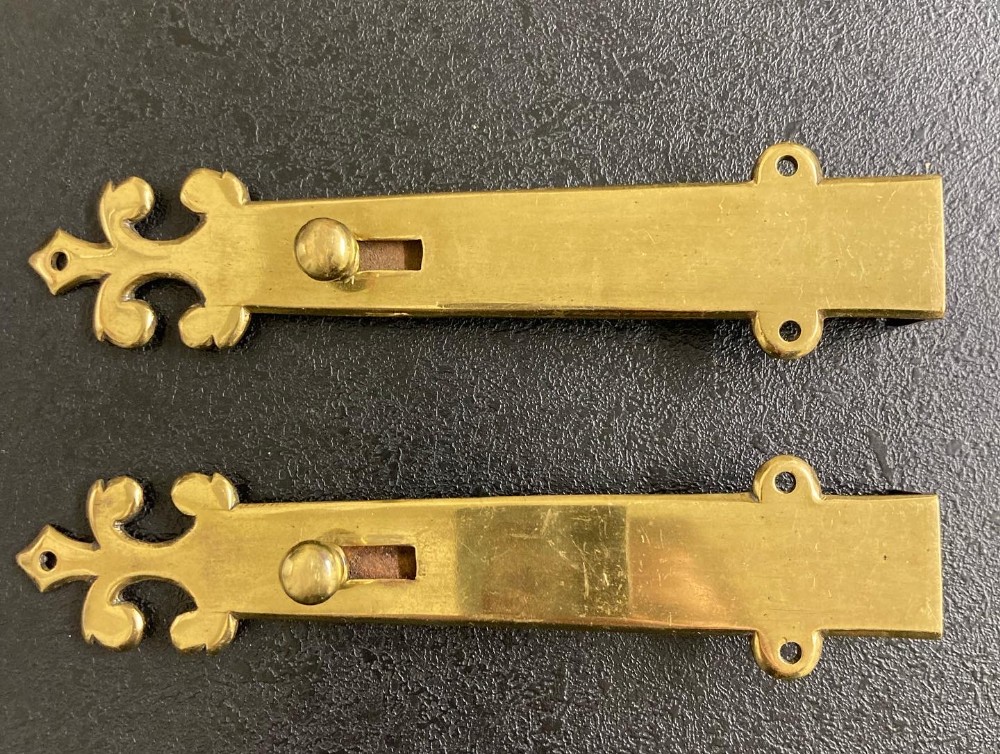 A matching pair of solid brass Victorian Gothic Revival surface-mounted door bolts featuring decorative fleur-de-lis terminals and round slider knobs.