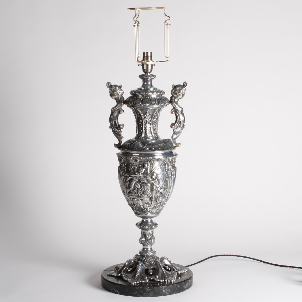 A polished silver-toned spelter table lamp base in the shape of a classical urn with relief carvings and creature handles on a circular marble plinth.