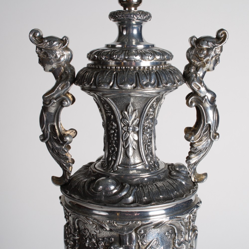 A polished silver-toned spelter table lamp base in the shape of a classical urn with relief carvings and creature handles on a circular marble plinth. View IV