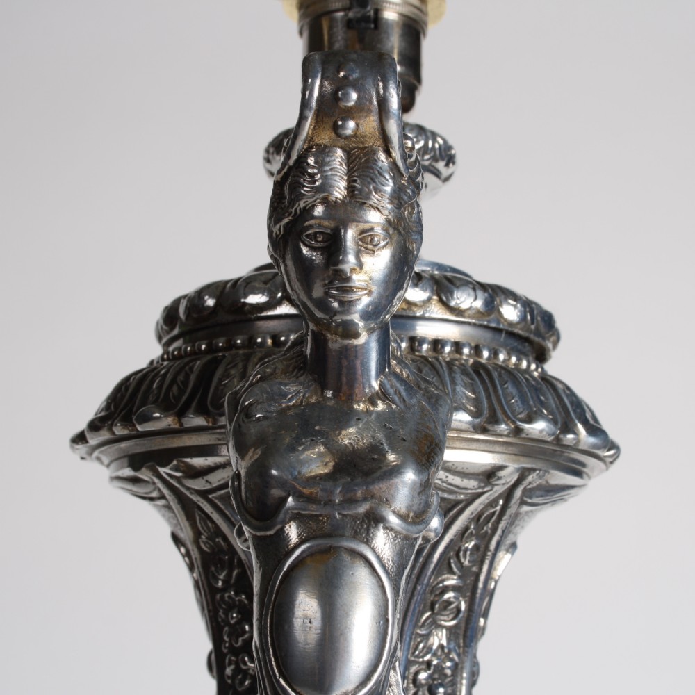 A polished silver-toned spelter table lamp base in the shape of a classical urn with relief carvings and creature handles on a circular marble plinth. View X