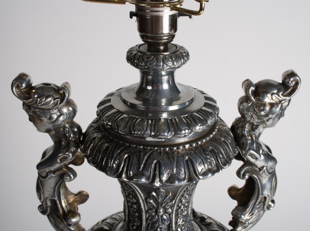 A polished silver-toned spelter table lamp base in the shape of a classical urn with relief carvings and creature handles on a circular marble plinth. View XII