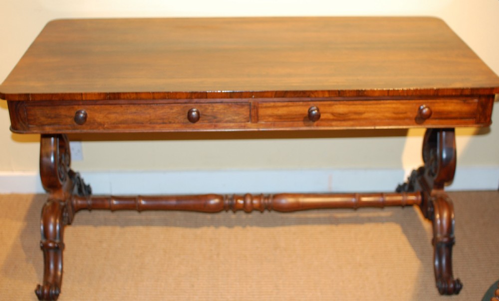 An antique rosewood library table featuring two frieze drawers with round timber knobs, supported by carved scroll trestle legs connected by a central turned stretcher.