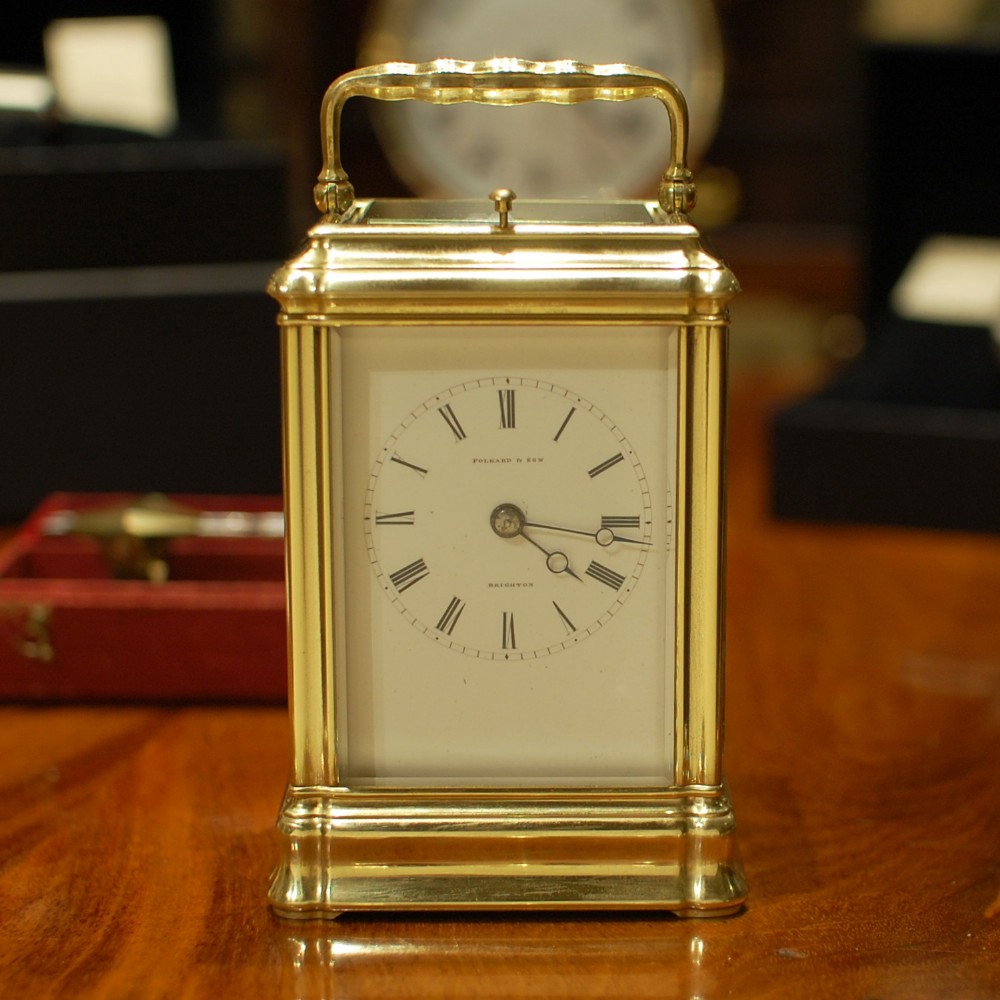 An antique brass carriage clock featuring a top carrying handle, glass viewing panels, and a white enamel dial with Roman numerals marked Folkard and Son Brighton.