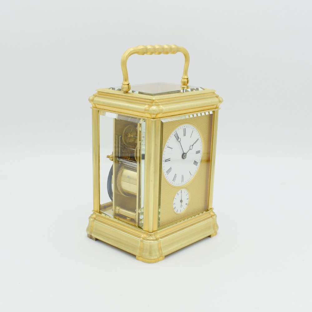 A gilt brass rectangular carriage clock featuring bevelled glass panels, a white enamel main dial with Roman numerals, a small subsidiary alarm dial, and a hinged carrying handle. View V