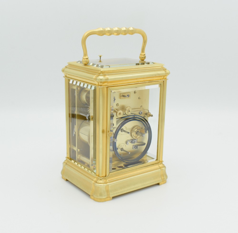 A gilt brass rectangular carriage clock featuring bevelled glass panels, a white enamel main dial with Roman numerals, a small subsidiary alarm dial, and a hinged carrying handle. View III