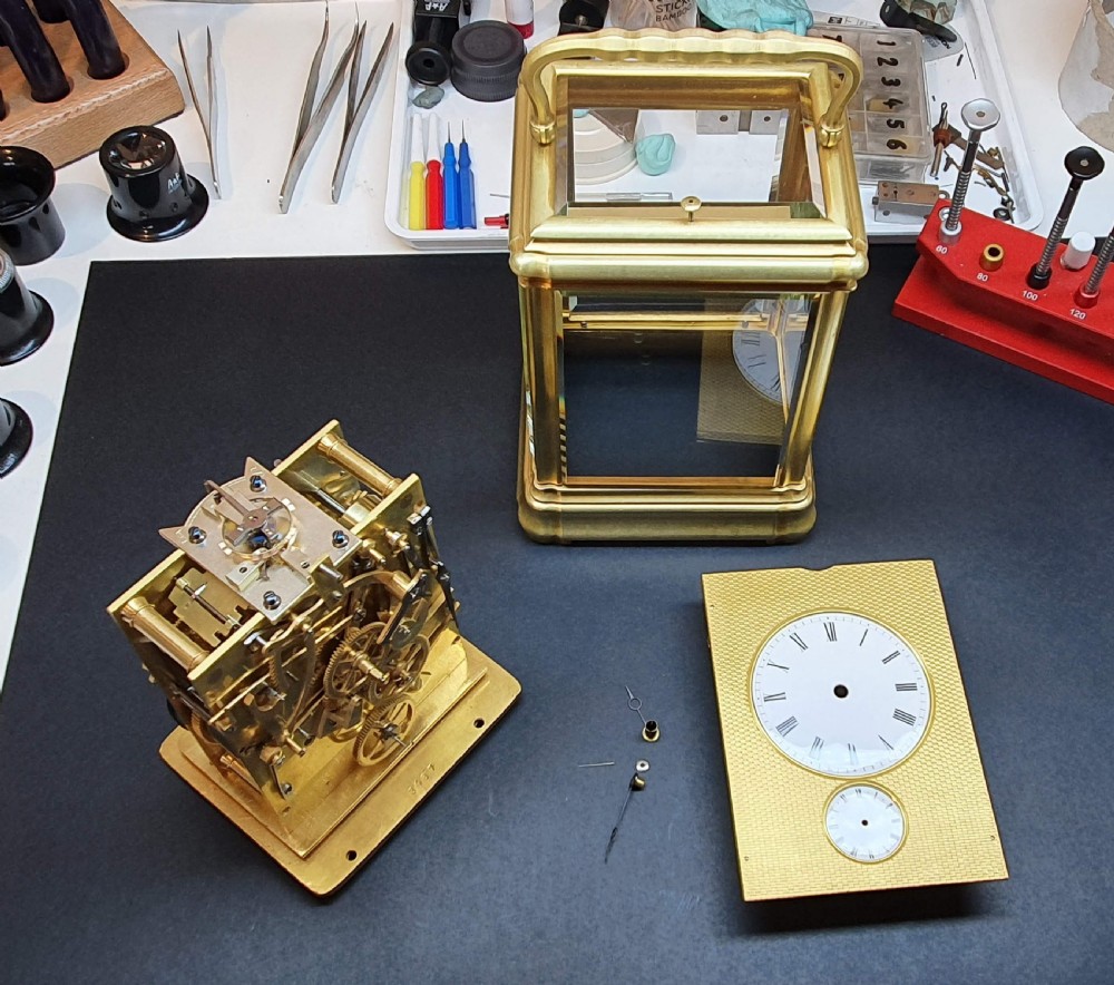 A gilt brass rectangular carriage clock featuring bevelled glass panels, a white enamel main dial with Roman numerals, a small subsidiary alarm dial, and a hinged carrying handle. View VII