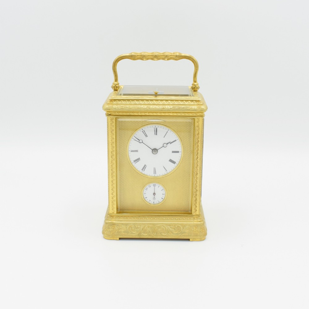 An antique gilded brass carriage clock featuring an engraved case, white enamel dial with Roman numerals, and bevelled glass panels.