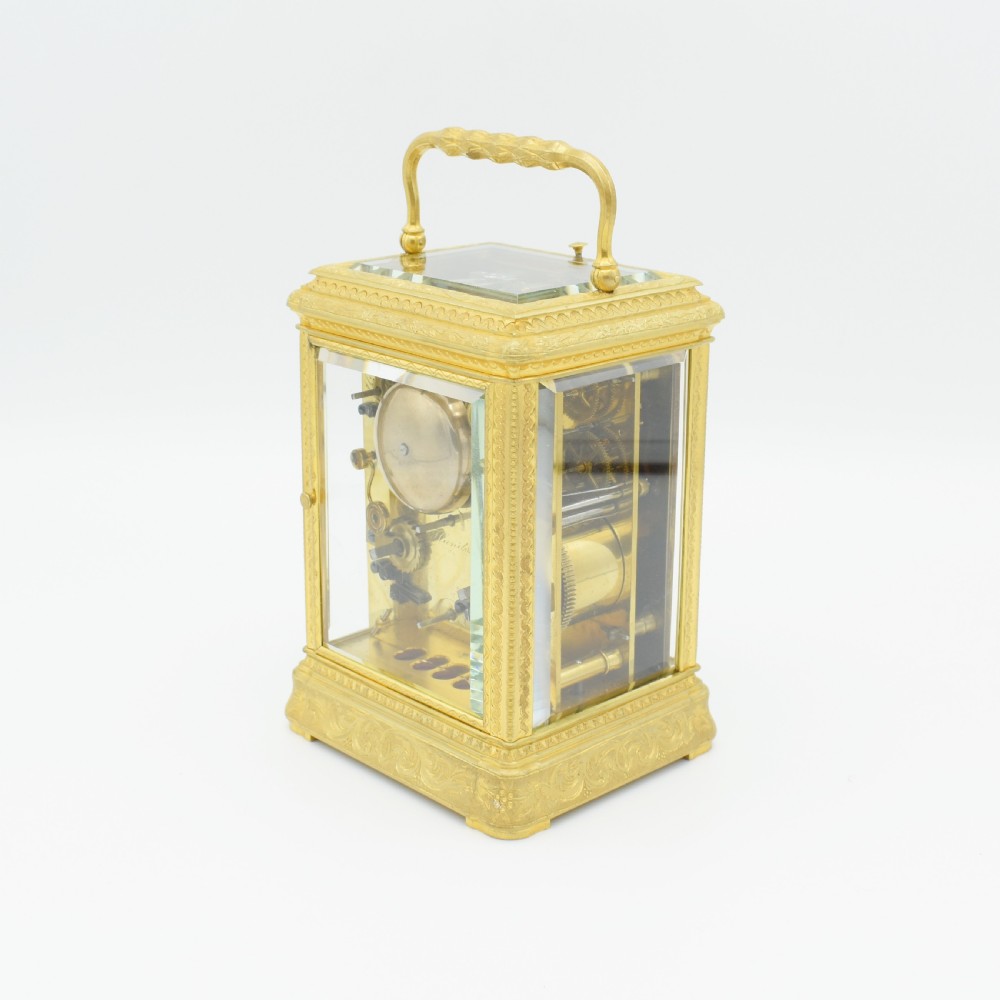 An antique gilded brass carriage clock featuring an engraved case, white enamel dial with Roman numerals, and bevelled glass panels. View IV