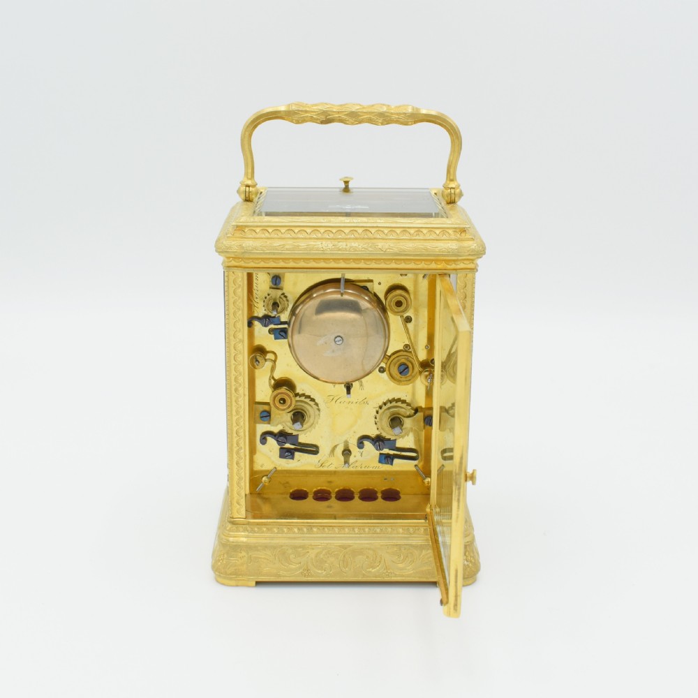 An antique gilded brass carriage clock featuring an engraved case, white enamel dial with Roman numerals, and bevelled glass panels. View V