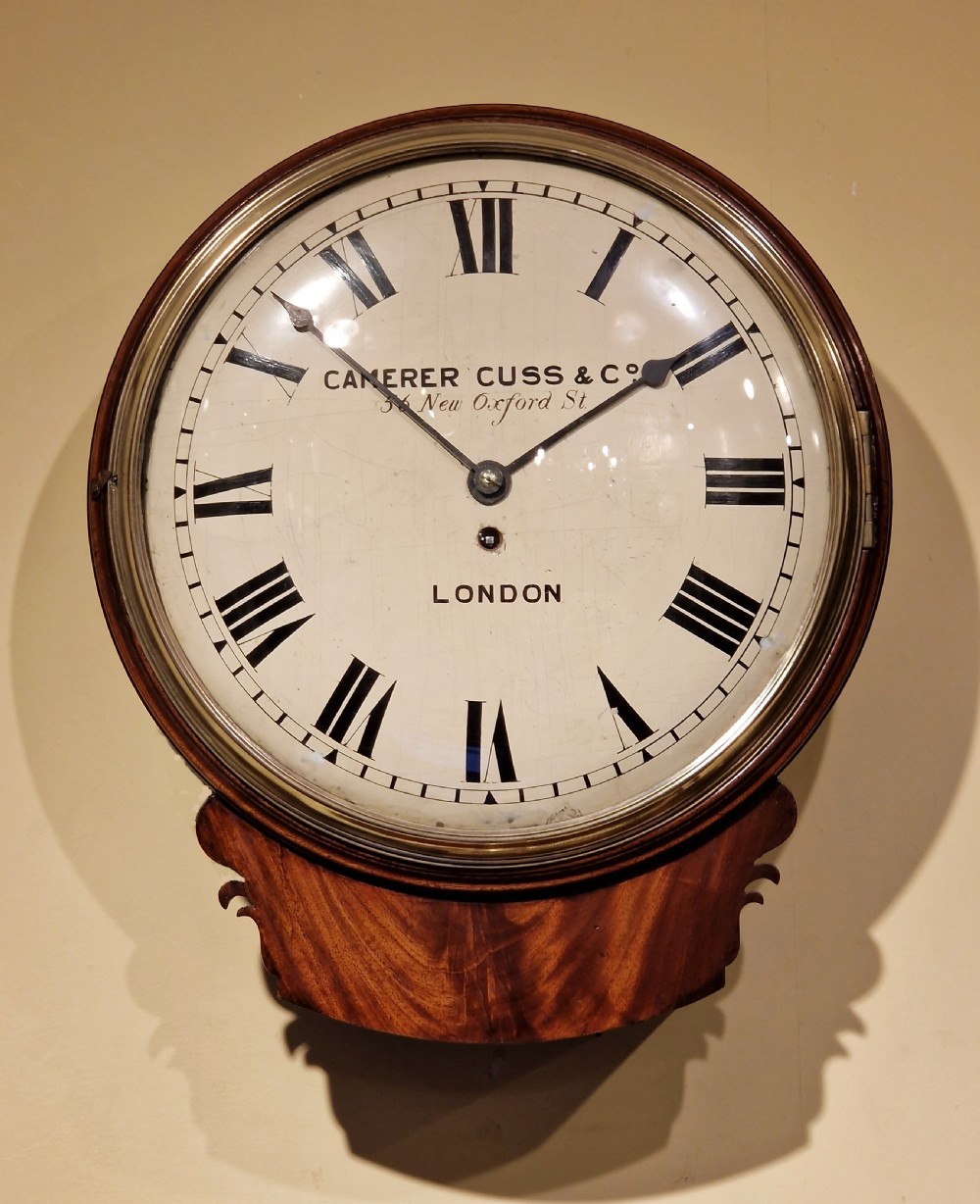 A large circular antique mahogany wall clock featuring a white painted dial with black Roman numerals, signed Camerer Cuss London, encased in a polished brass rimmed bezel with a lower wood bracket.