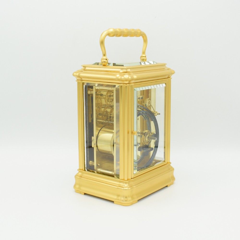 A gilded brass carriage clock featuring a shaped top carrying handle, clear bevelled glass panels, and a white enamel dial with Roman numerals. View VI