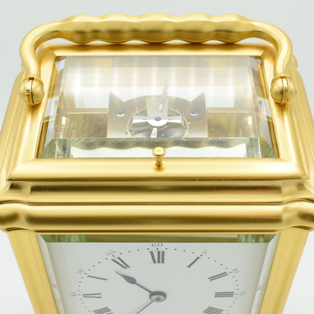 A gilded brass carriage clock featuring a shaped top carrying handle, clear bevelled glass panels, and a white enamel dial with Roman numerals. View VII