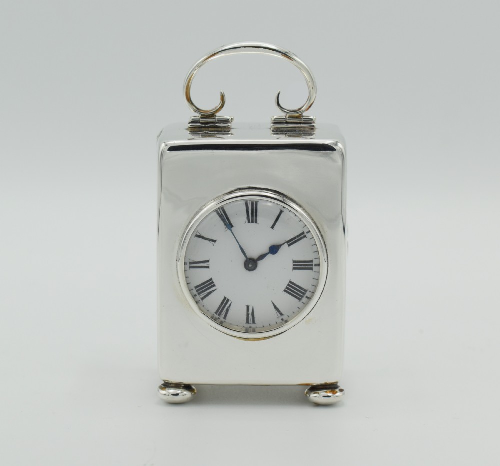 A small rectangular polished silver carriage clock featuring a top carrying handle, white enamel dial with Roman numerals, and bun feet.