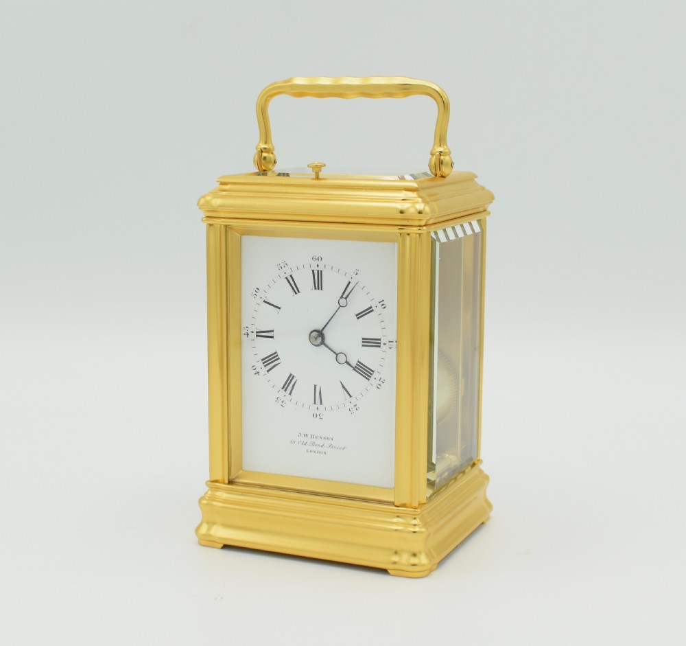 A rectangular brass carriage clock featuring bevelled glass side panels, a white enamel dial with Roman numerals, and a hinged handle on top.