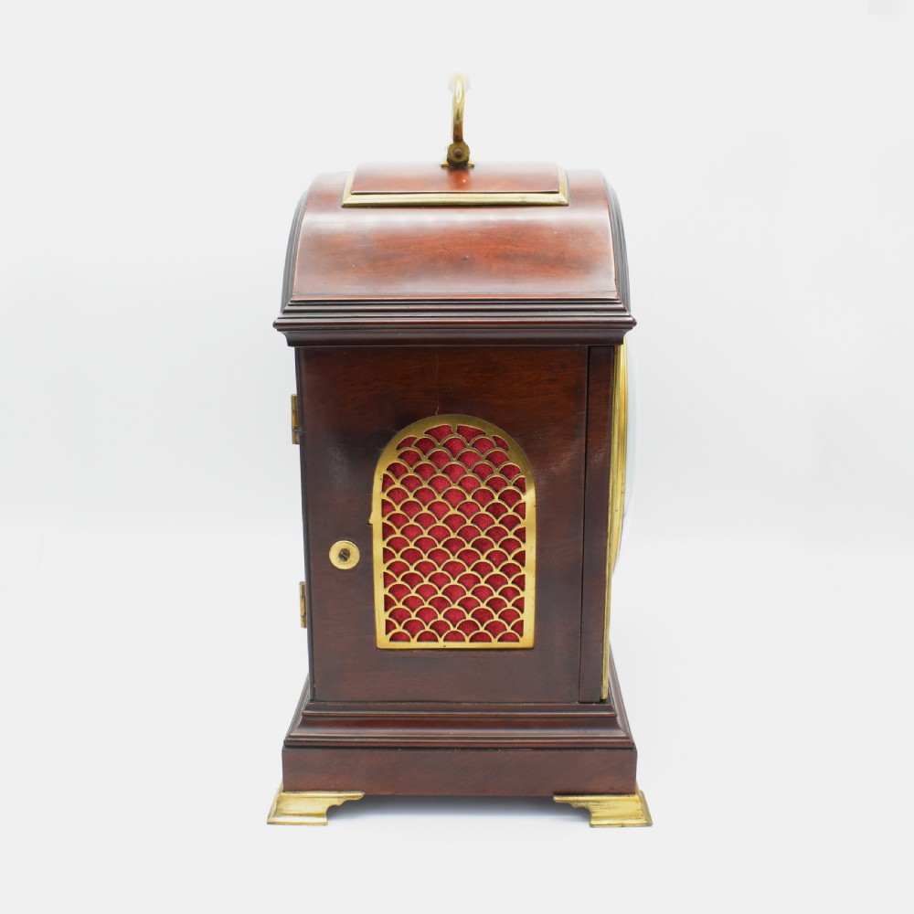 An antique mahogany bracket clock featuring an arched top, brass carrying handle, white dial with Roman numerals, and brass fretwork spandrels backed with red silk. View III