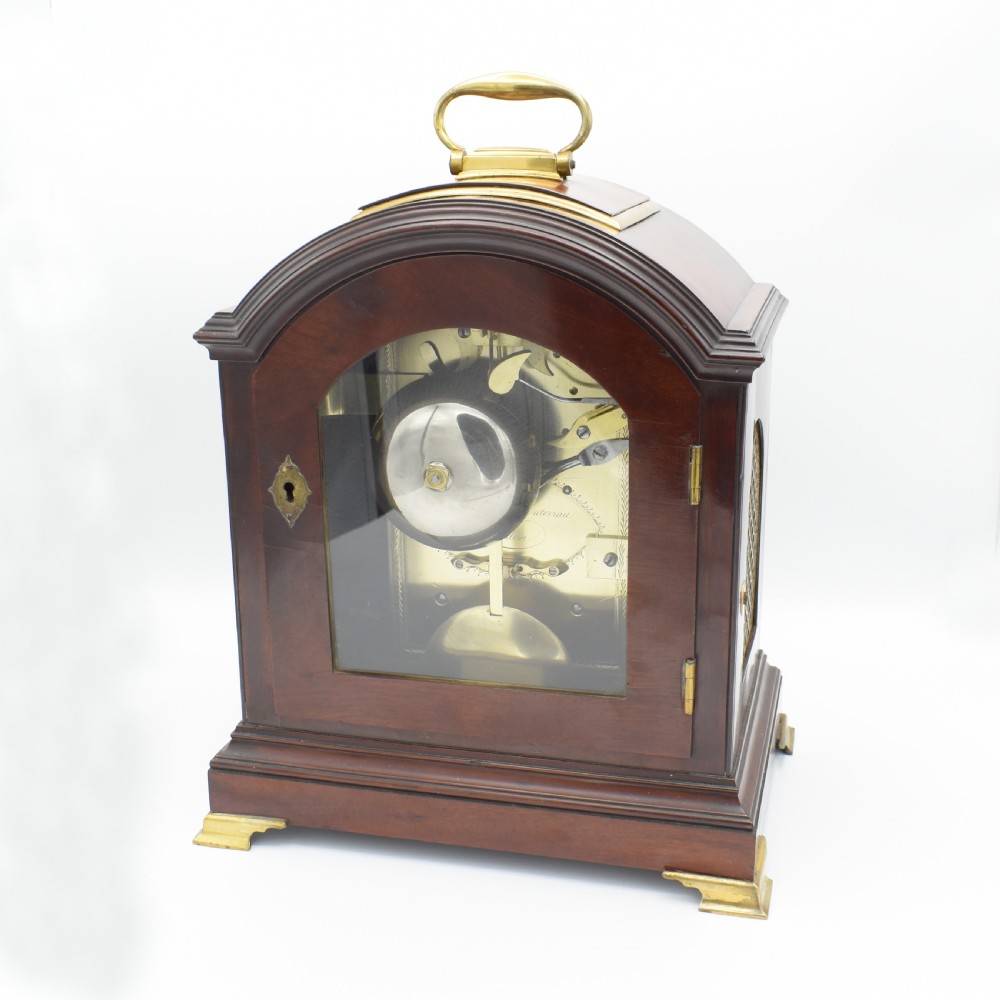 An antique mahogany bracket clock featuring an arched top, brass carrying handle, white dial with Roman numerals, and brass fretwork spandrels backed with red silk. View IV