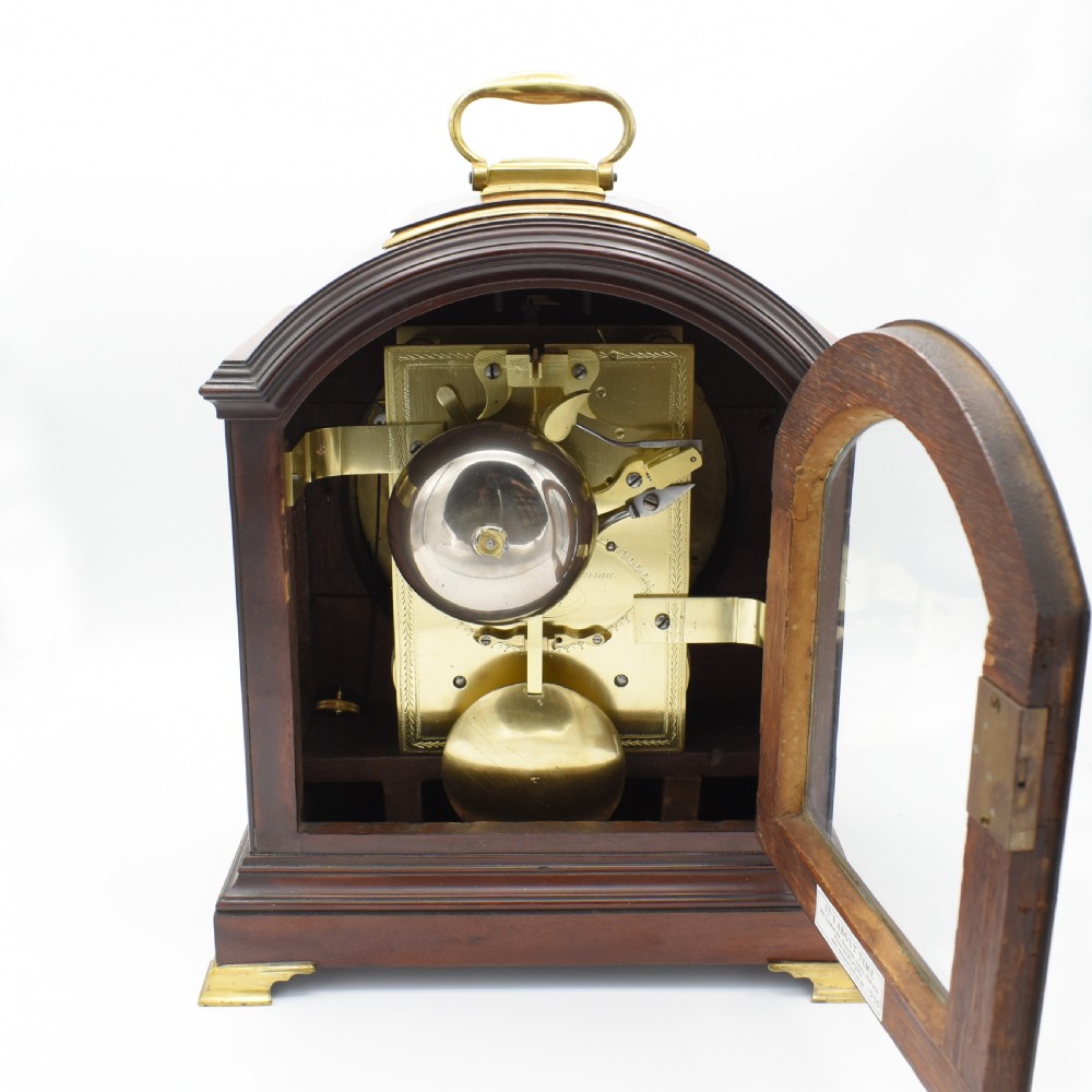 An antique mahogany bracket clock featuring an arched top, brass carrying handle, white dial with Roman numerals, and brass fretwork spandrels backed with red silk. View V