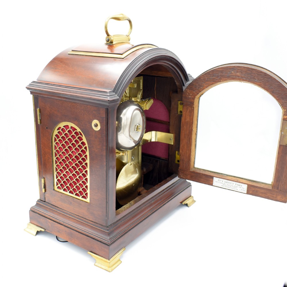An antique mahogany bracket clock featuring an arched top, brass carrying handle, white dial with Roman numerals, and brass fretwork spandrels backed with red silk. View VII
