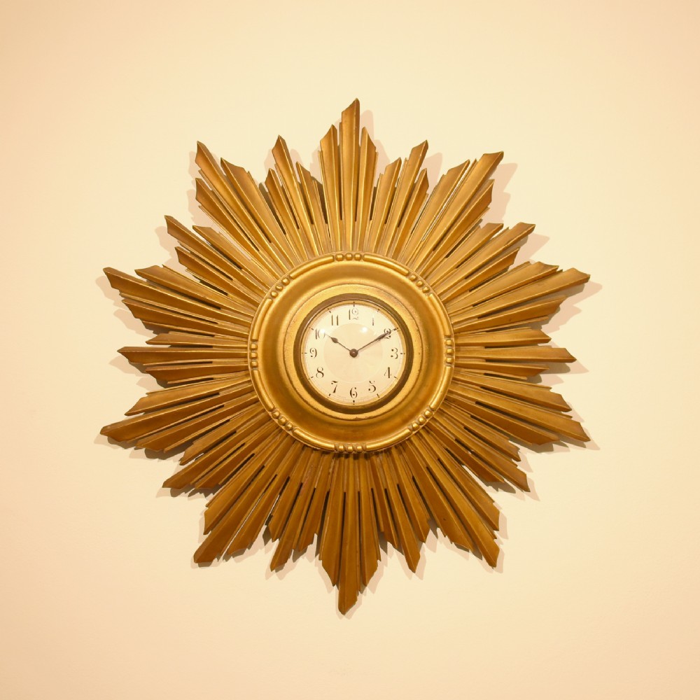 A circular giltwood sunburst wall clock featuring layered radiating wooden rays surrounding a central white dial with Arabic numerals.