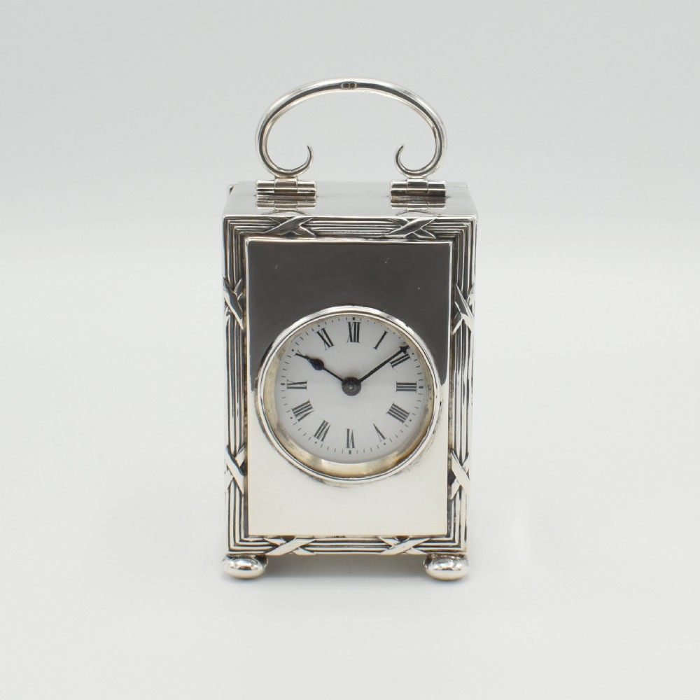A rectangular sterling silver carriage clock featuring a top looped handle, white enamel dial with black Roman numerals, and decorative reeded borders resting on bun feet.