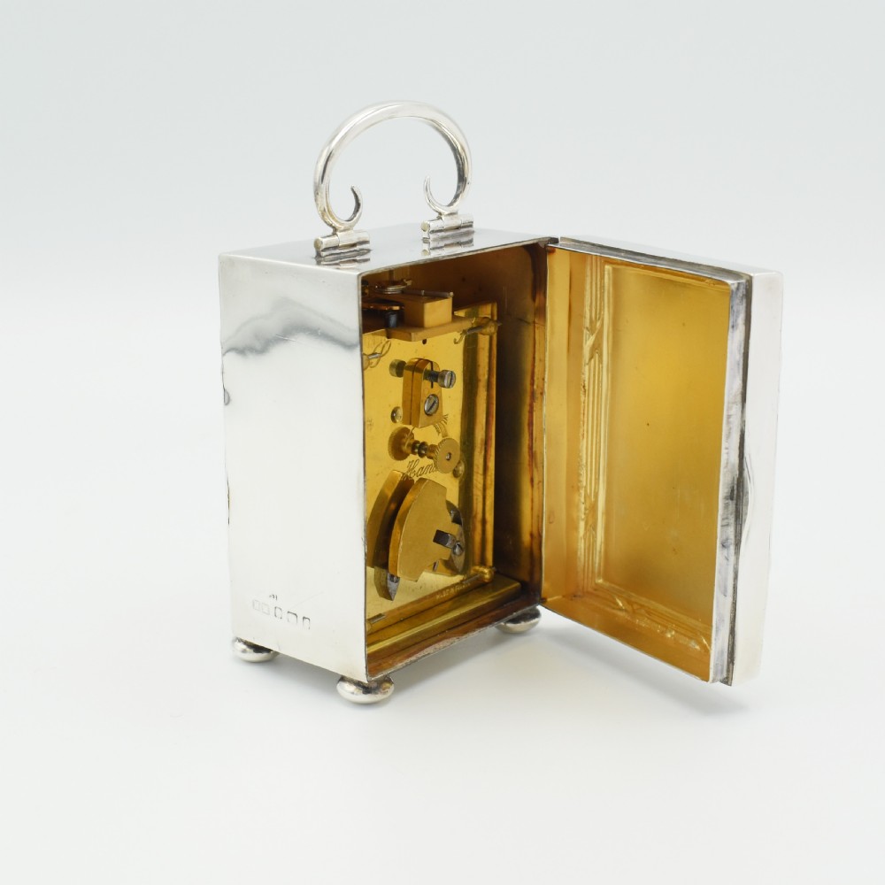 A rectangular sterling silver carriage clock featuring a top looped handle, white enamel dial with black Roman numerals, and decorative reeded borders resting on bun feet. View II