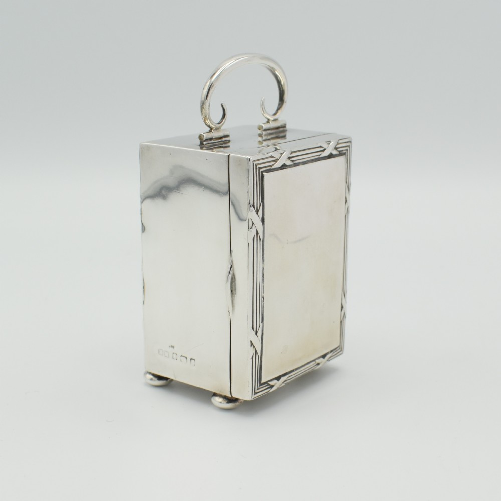 A rectangular sterling silver carriage clock featuring a top looped handle, white enamel dial with black Roman numerals, and decorative reeded borders resting on bun feet. View III
