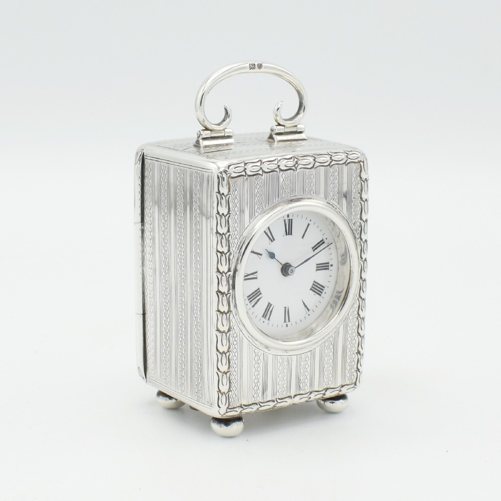 A miniature sterling silver carriage clock featuring engine-turned engraving, a white enamel dial with Roman numerals, a top carrying handle, and ball feet.