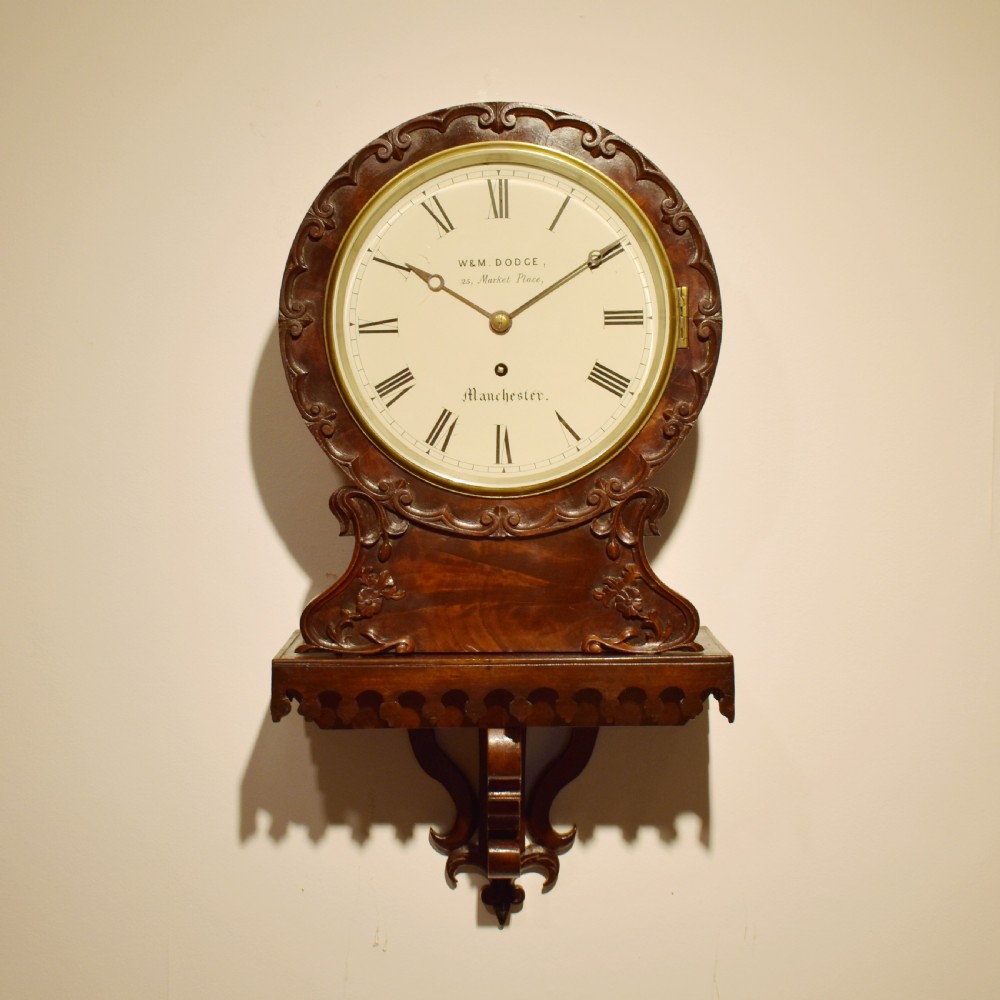 A small Victorian mahogany wall clock featuring a circular white dial with Roman numerals, a brass bezel, and carved foliate detailing on an integrated bracket base.
