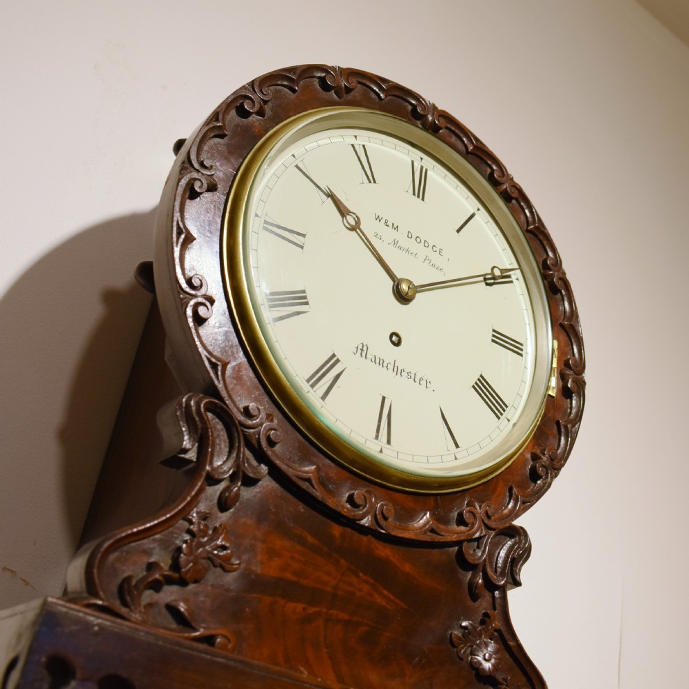 A small Victorian mahogany wall clock featuring a circular white dial with Roman numerals, a brass bezel, and carved foliate detailing on an integrated bracket base. View II