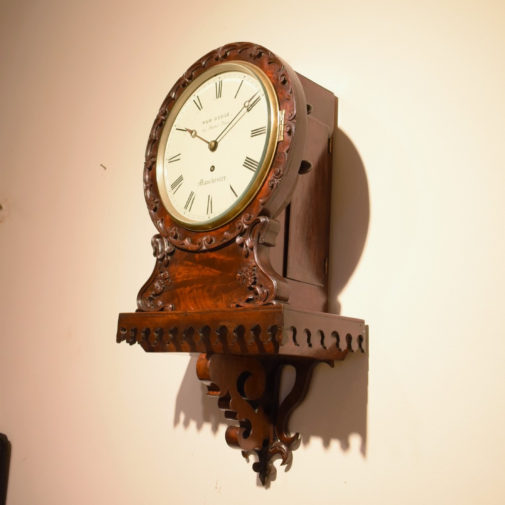 A small Victorian mahogany wall clock featuring a circular white dial with Roman numerals, a brass bezel, and carved foliate detailing on an integrated bracket base. View III