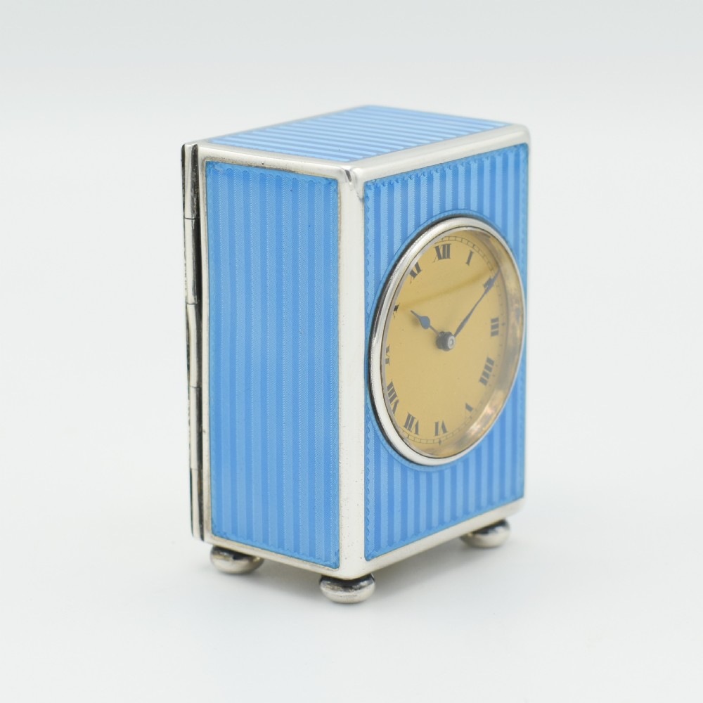 An Art Deco rectangular miniature clock featuring sky blue guilloche enamel casing with a circular dial, Roman numerals, and silver ball feet. View IV