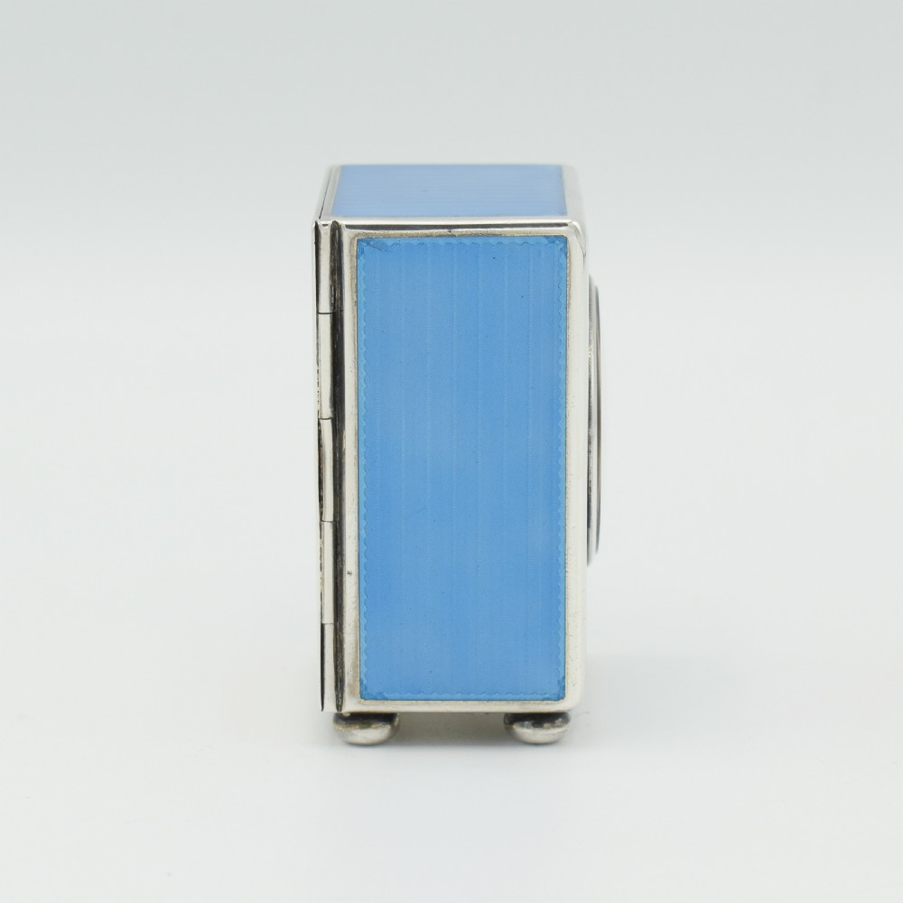 An Art Deco rectangular miniature clock featuring sky blue guilloche enamel casing with a circular dial, Roman numerals, and silver ball feet. View VIII