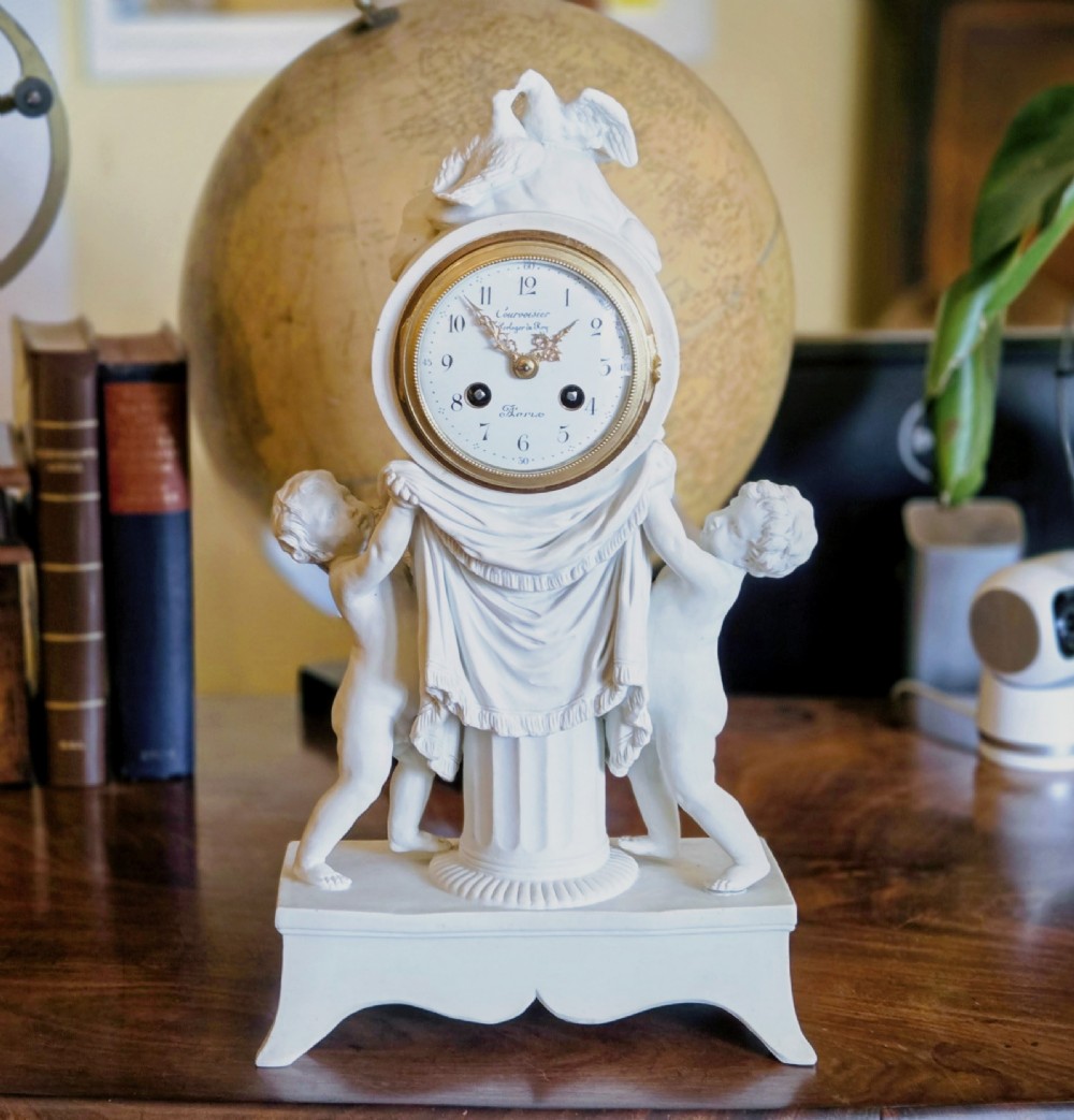 A white bisque porcelain Neoclassical mantel clock featuring two putti flanking a fluted column topped with a pair of doves.