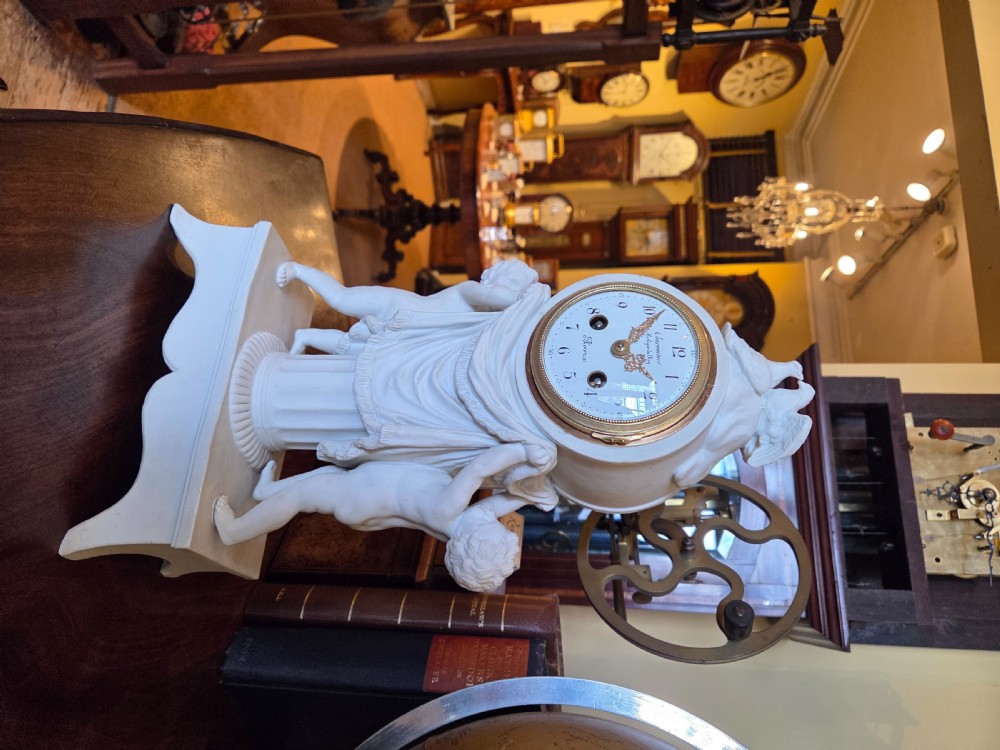 A white bisque porcelain Neoclassical mantel clock featuring two putti flanking a fluted column topped with a pair of doves. View II