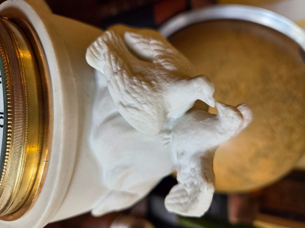 A white bisque porcelain Neoclassical mantel clock featuring two putti flanking a fluted column topped with a pair of doves. View IV