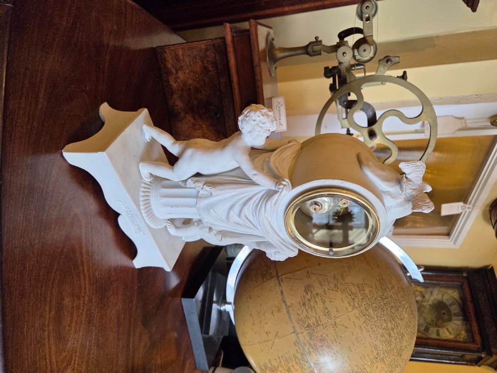 A white bisque porcelain Neoclassical mantel clock featuring two putti flanking a fluted column topped with a pair of doves. View V