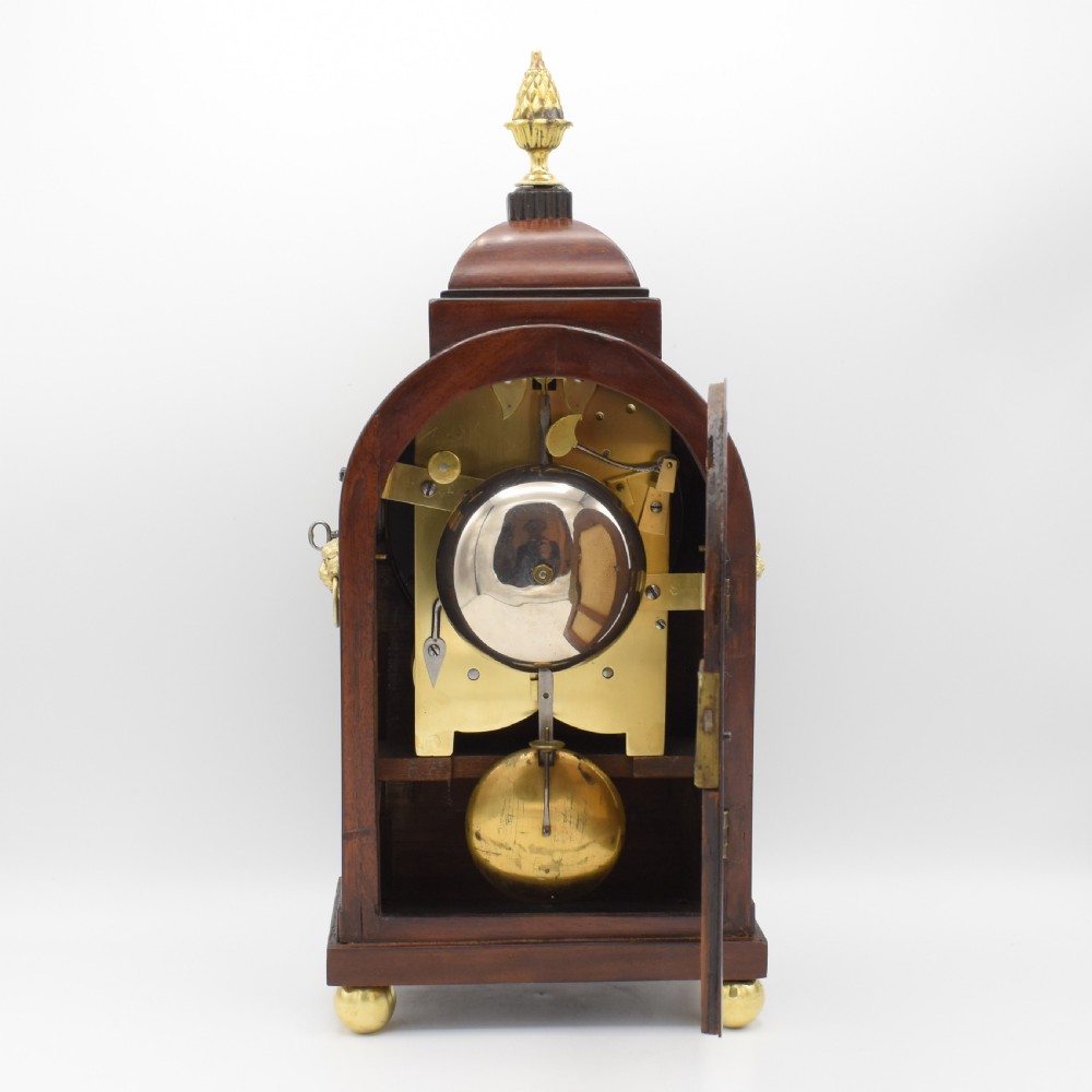 An antique mahogany bracket clock featuring a brass pineapple finial, brass inlay details, a cream dial with Roman numerals, and brass ball feet. View V