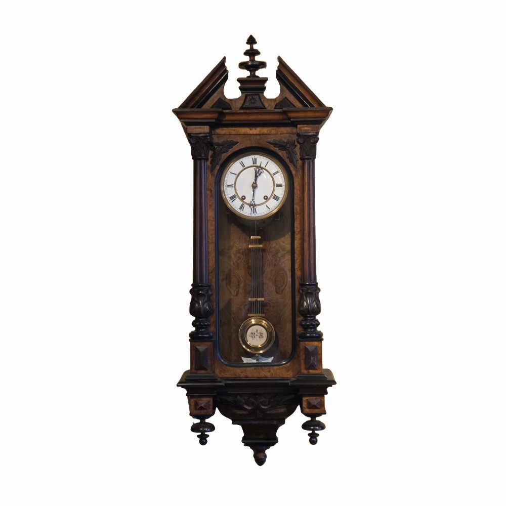 A late nineteenth-century German Vienna regulator wall clock in carved dark walnut with turned columns, glass door, white enamel dial with Roman numerals, and visible pendulum.