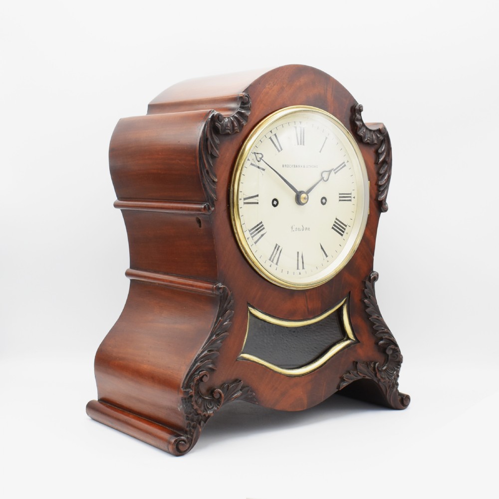 An antique mahogany bracket clock with carved acanthus scrollwork, a cream dial featuring Roman numerals, and a glass aperture revealing the pendulum. View II