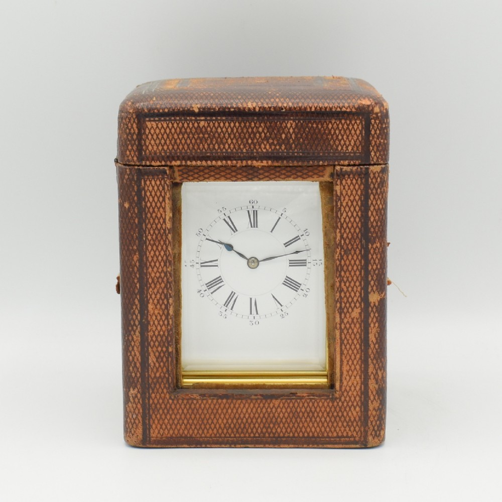 An antique French brass carriage clock featuring a white enamel dial with Roman numerals, set within a textured brown leather travel case.