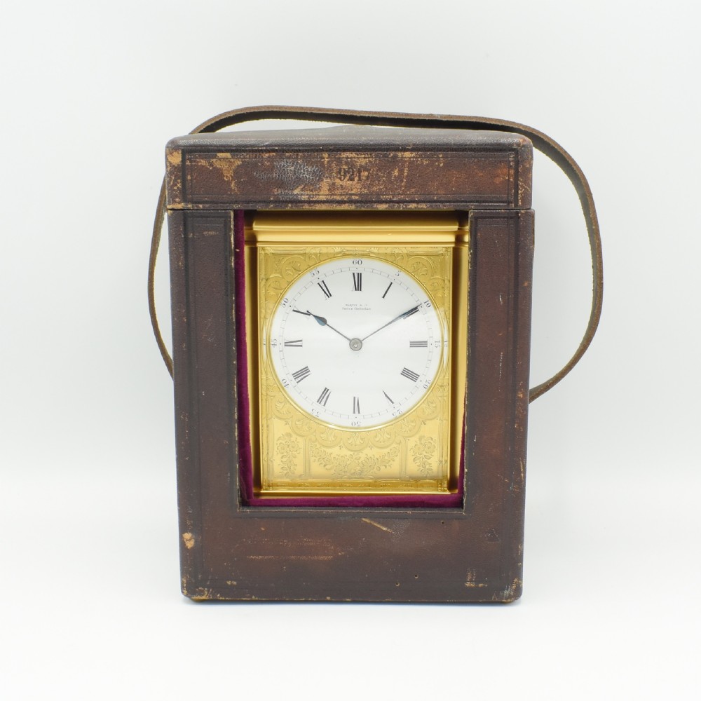 An antique gilt brass carriage clock with an engraved face and white enamel dial housed inside a fitted dark brown leather travelling case with a handle.
