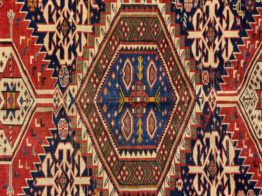 An antique hand-knotted Caucasian Shirvan wool rug featuring central geometric diamond medallions and intricate layered borders in red, cream, and navy blue. View III