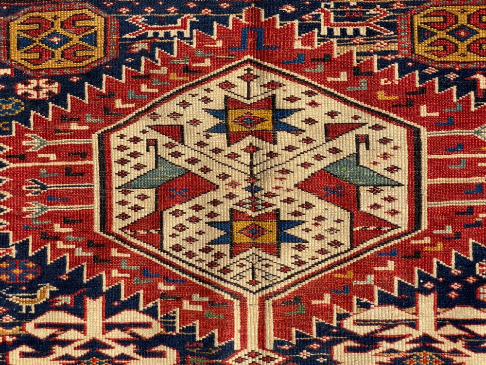 An antique hand-knotted Caucasian Shirvan wool rug featuring central geometric diamond medallions and intricate layered borders in red, cream, and navy blue. View IV