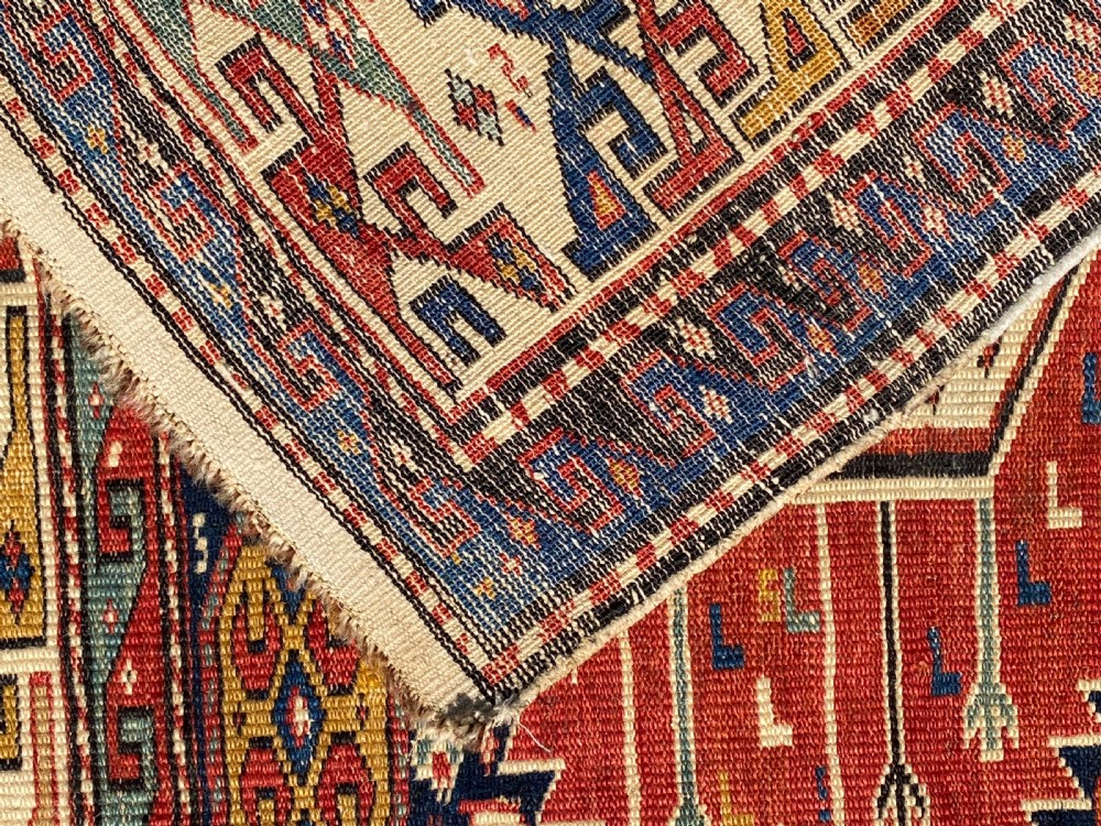 An antique hand-knotted Caucasian Shirvan wool rug featuring central geometric diamond medallions and intricate layered borders in red, cream, and navy blue. View X