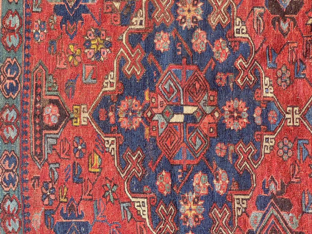 An antique Caucasian Soumak wool rug featuring geometric stepped medallions on a terracotta red field with blue and multi-coloured borders. View III