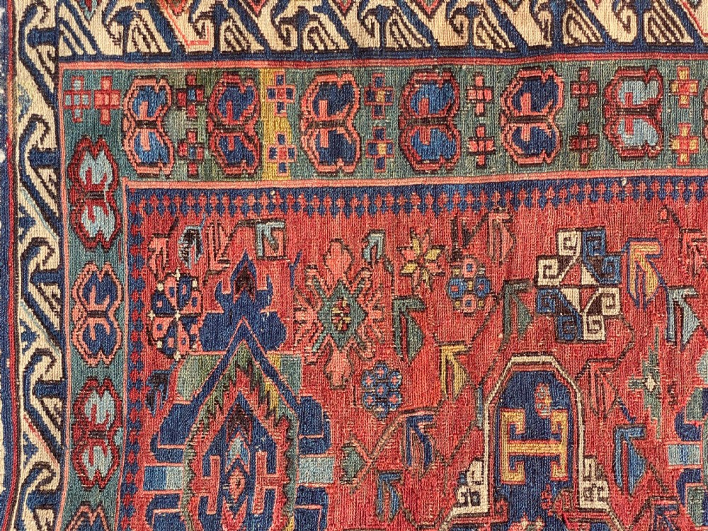 An antique Caucasian Soumak wool rug featuring geometric stepped medallions on a terracotta red field with blue and multi-coloured borders. View IV