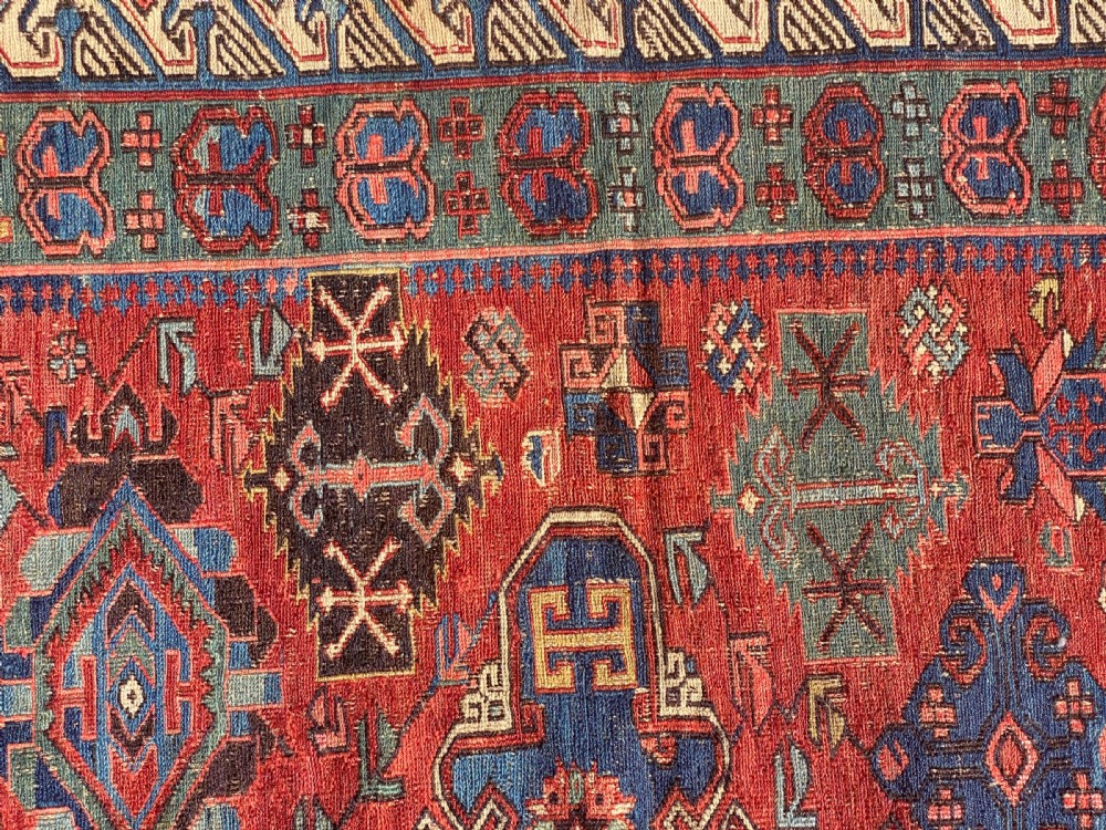 An antique Caucasian Soumak wool rug featuring geometric stepped medallions on a terracotta red field with blue and multi-coloured borders. View V